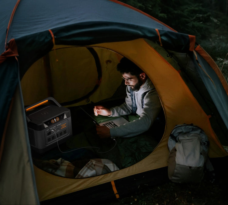 A man sits inside a camping tent at night, using a laptop and charging a portable power station. A backpack rests beside him on the tent floor, with a dark forest background visible outside the tent.