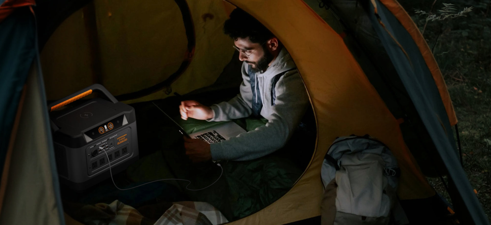 A man sits inside a camping tent at night, using a portable power station and laptop. The tent is illuminated by warm light, with a backpack visible on the floor and a forest setting outside.